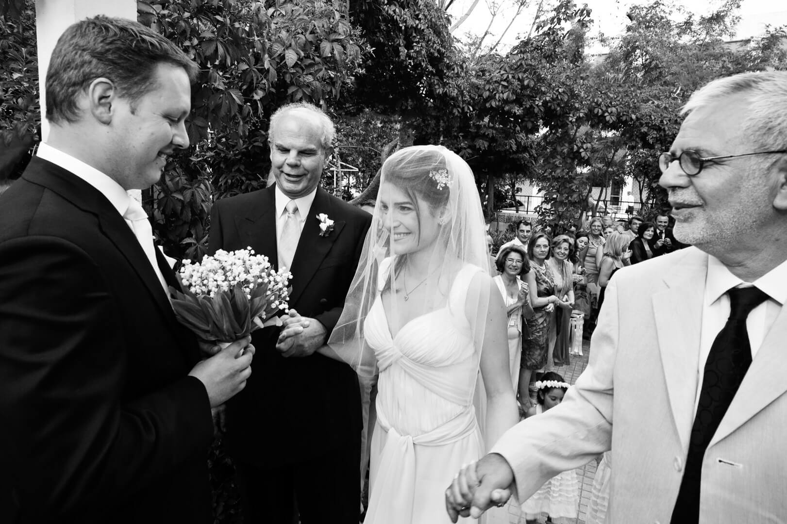 Bride radiates joy, flanked by father and stepfather, as groom presents her with a beautiful bouquet at the church entrance.