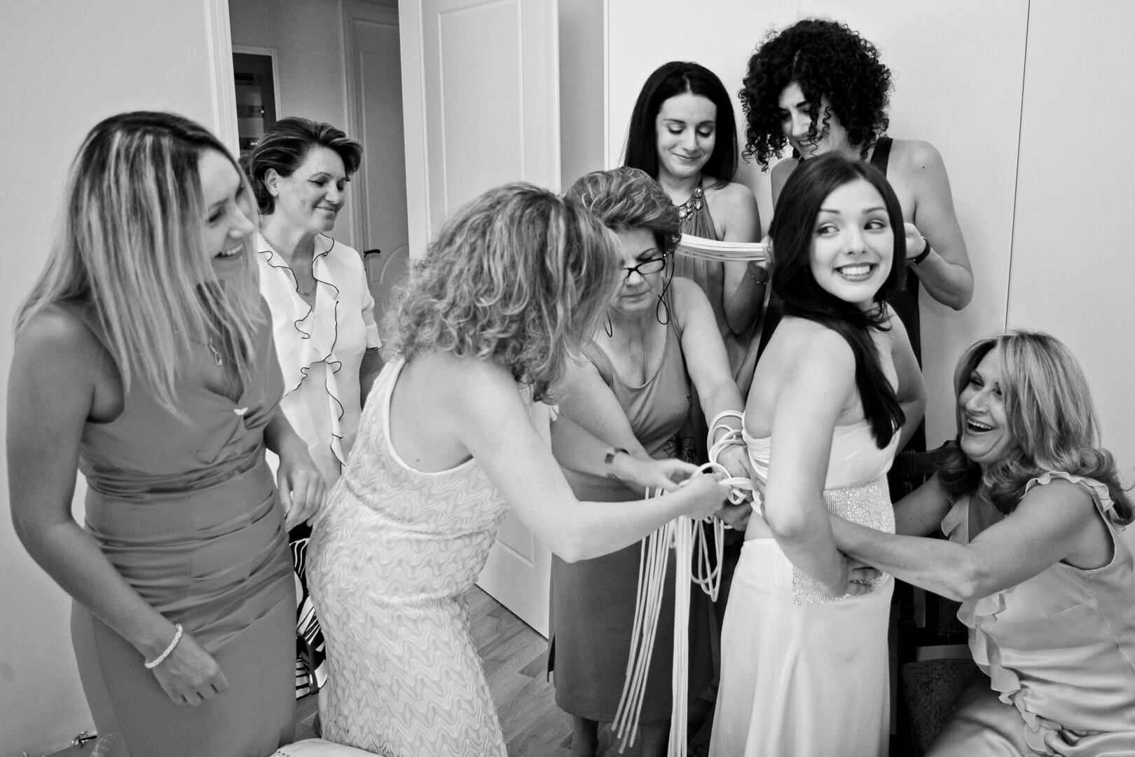 Bride joyfully assisted by friends and family, tying up her wedding dress with love and care.