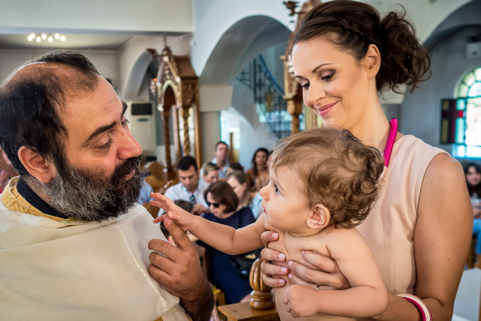 Precious interaction during baptism: priest offers blessings while a curious child reaches out. 