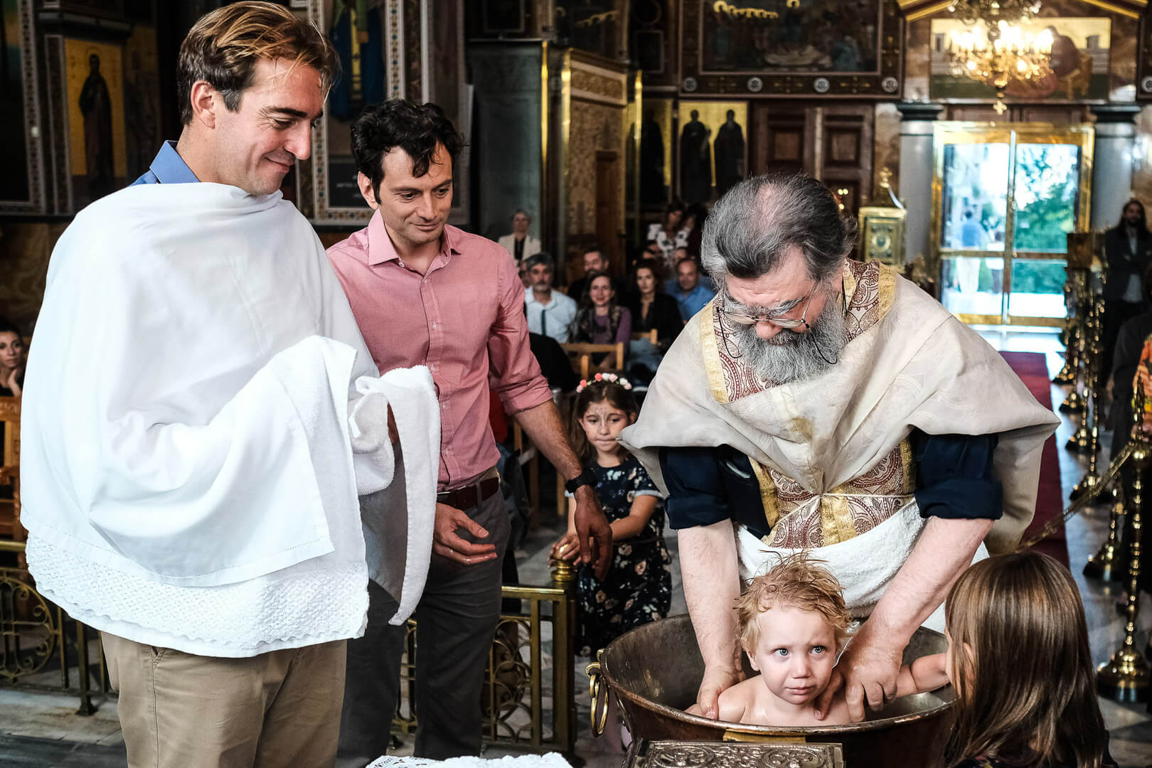 Greek christening traditions: two godfathers stand by as baby receives blessings at baptism.
